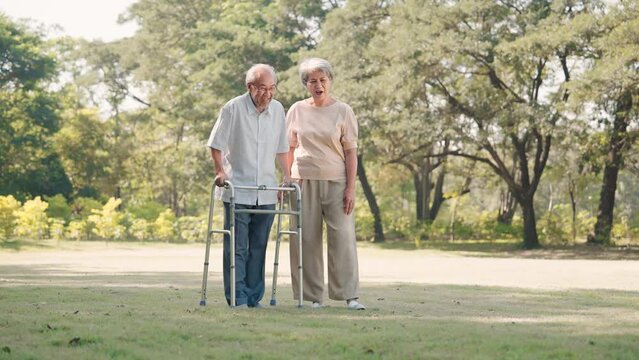 Asian senior woman prop up husband walking with walker cane in the public park, Physical therapy concept