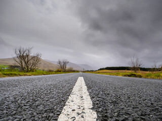 Small narrow asphalt road to mountains in low cloudy sky. Travel background. Calm Irish nature landscape. Transportation and tourism concept. West of Ireland.