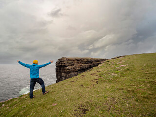 Obraz premium Male tourist in blue jacket and yellow hat standing on edge of a cliff, Loop head area county Clare, Ireland. Dramatic sky. Travel and sightseeing