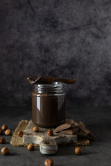 Obraz na płótnie Canvas Wooden spoon with chocolate paste on a transparent jar. Hazelnuts and pieces of chocolate on the table. Dark background, space for text