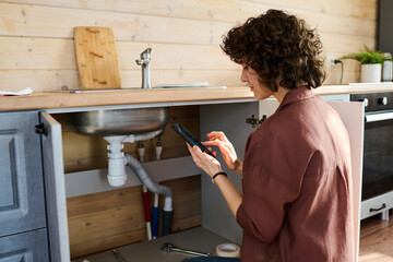 Young brunette housewife with mobile phone squatting by open cabinet of kitchen counter while...