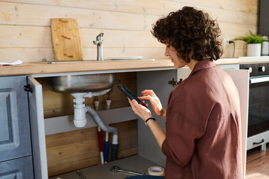 Young brunette housewife with mobile phone squatting by open cabinet of kitchen counter while sending photo of damaged pipe to plumber