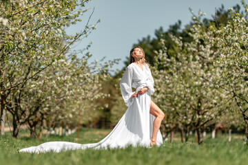 Fototapeta premium Blond blooming garden. A woman in a white dress walks through a blossoming cherry orchard. Long dress flies to the sides,