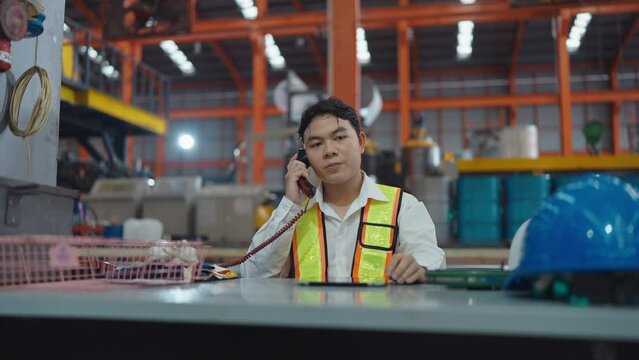Young asian warehouse worker in safety uniform answer the phone talking with manager using tablet while working at industrial warehouse