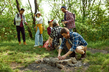 Group of friends, young men and women going hiking in forest, camping, doing picnic on warm summer day. Boy setting a fire. Concept of active lifestyle, nature, sport and hobby, friendship