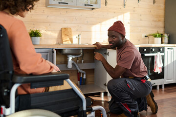 Fototapeta premium Young repairman pointing at pipe under kitchen sink and looking at housewife in wheelchair while explaining her the problem