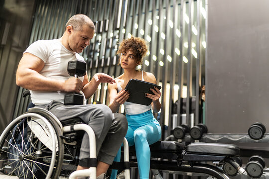 A determined individual in a wheelchair, working out in the gym with the guidance of a dedicated female fitness trainer who using a tablet to show a weekly workout plan.