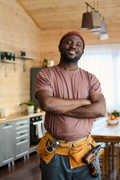 Young successful electrician or fitterman with toolbelt crossing arms by chest and looking at camera while standing in spacious kitchen