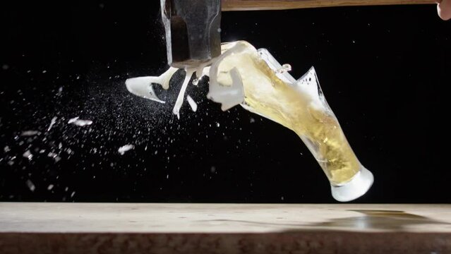 Beer glass smashed with sledge hammer spills beer everywhere, black background