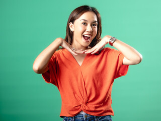 Fototapeta premium A studio portrait of a beautiful young Asian Indonesian woman wearing an orange shirt her hand pointing towards her mouth . Isolated on a green background.