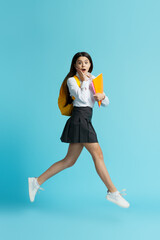 Portrait of shocked girl wearing school uniform, backpack, holding notebooks jumping high isolated on blue background. Back to school, education concept