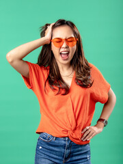 A portrait of an Asian woman wearing an orange shirt and glasses, feeling dizzy and holding her head with one hand, isolated on a green background.