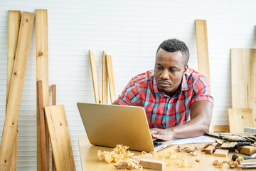 Young african american male carpenter sitting at table working with tablet in workshop, craftsman constructing wooden home model by learning from online