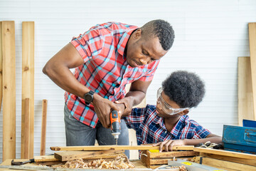 Careful father is teaching his son to work with electric screwdriver while son is trying to use screw gun and fix screw in pieces of wood. Early childhood education concept