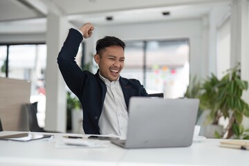 Happy young businessman looking at laptop excited by good news online, lucky successful winner man sitting at office desk raising hand in yes gesture celebrating business success win result
