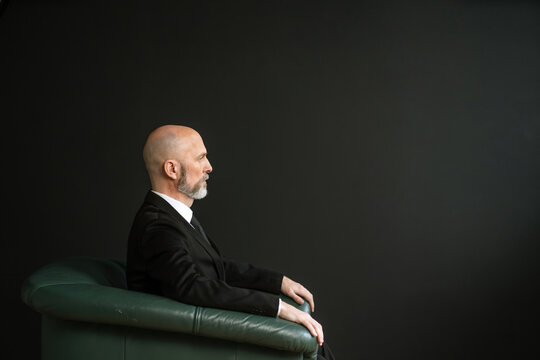 Mature man in a suit, sitting in a comfortable leather chair. The backdrop of a black wall adds a touch of elegance and sophistication to the image.