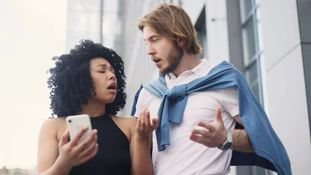 Portrait of amazed cute interracial couple got notification message and looking at camera with puzzled face outdoors Shocked multiethnic people reacting to news and feel confused at city street