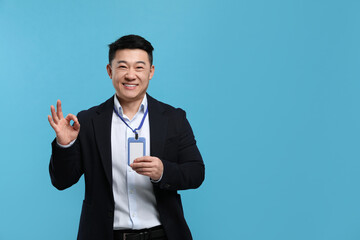 Happy asian man with vip pass badge showing ok gesture on light blue background. Space for text