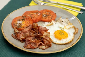 Closeup of an a fried egg, bacon and tomatoes on a gray plate laid on a green tablecloth