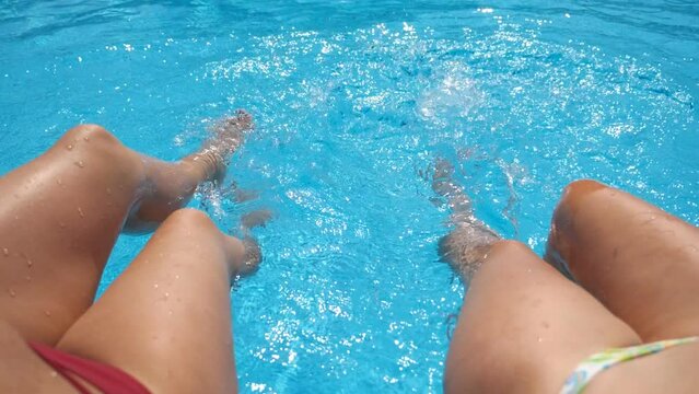 Female legs splashing crystal clear water in pool of hotel. Two friends sitting on edge of basin and enjoying their vacation at sunny day. Concept of summer vacation or holiday. Slow mo Point of view