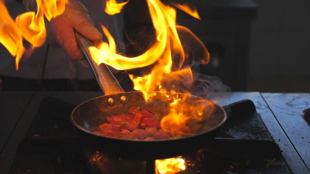 Detail view on flaming skillet in hand of professional chef. Male cook mixing and frying vegetables cooking tasty flambe style dish for visitors of restaurant. Concept of cooking. Slow mo Front view