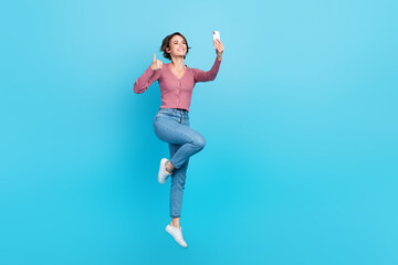 Fototapeta na wymiar Full body photo of charming carefree lady jumping hold smartphone show thumb up isolated on blue color background