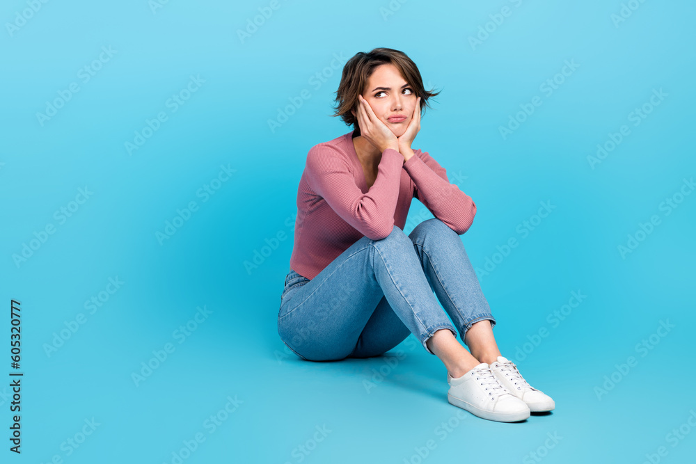 Poster Full size photo of unsatisfied minded girl sit floor hands touch cheeks look empty space isolated on blue color background