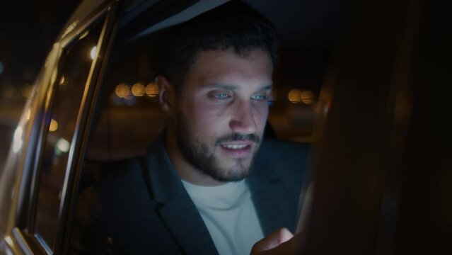 Close Up Slow Motion of Handsome Young Caucasian Man in Car Using Mobile Phone. Male Bearded Brunette on Backseat of Ride Sharing Transportation Checking Social Media Posts in Smartphone. Night 