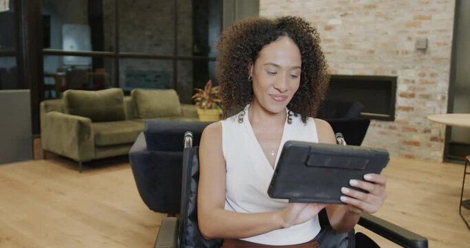 Portrait of biracial businesswoman in wheelchair using tablet in office and smiling, in slow motion