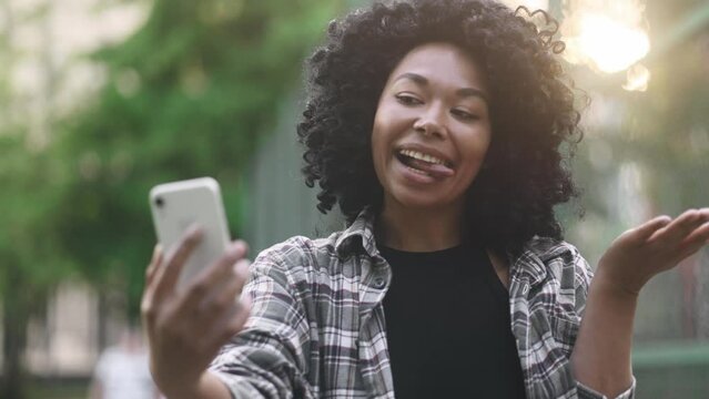 Portrait of young african american woman grimacing taking selfie photograph saving great memories on smartphone mobile phone at green street Pretty curly girl create content for social media outdoors