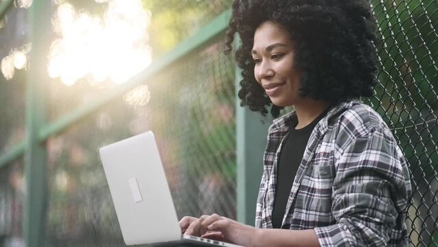 Pretty young african american woman freelancer or student working on laptop at green street Curly girl having distance remote freelance work or education on computer and looking at camera outdoors