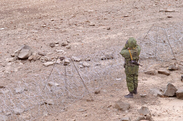 An armed soldier in patrol with binoculars guards an area bounded by a barbed wire fence....