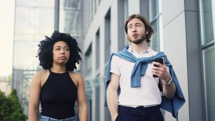 Narcissistic handsome young man gives an inspired introduction to his companion on date while she boring and rolls her eyes at city street Interracial people met after texting on dating app outdoors 