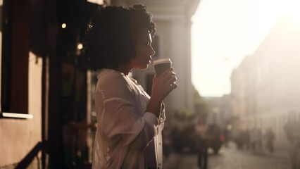 Cinematic shot of serene young African American curly woman hold cup enjoying aroma of hot coffee or tea with beautiful sun rays on her face at urban city street Beautiful day concept