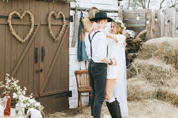 young couple newlyweds bride in short white dress and wreath with veil and groom in hat and jeans with suspenders hugging and having fun at barn of country house