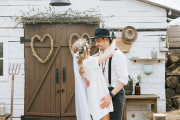young couple newlyweds bride in short white dress and wreath with veil and groom in hat and jeans with suspenders hugging and having fun at barn of country house © klavdiyav