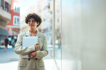Fototapeta na wymiar Portrait of cute confident mature adult business woman or manager standing near work desk in office, dressed in formal stylish clothes and glasses, holding laptop, looking at camera, smiling friendly