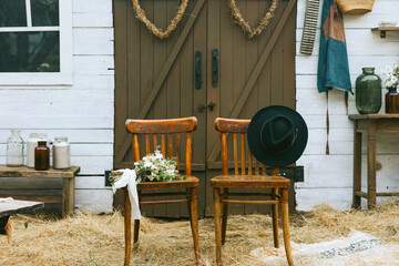 empty chairs with hat and bouquet of bride near barn of country house at boho style wedding celebration, boho decorations with macrame and hay