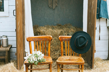 empty chairs with hat and bouquet of bride near barn of country house at boho style wedding...