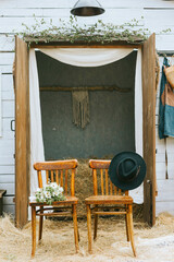 empty chairs with hat and bouquet of bride near barn of country house at boho style wedding celebration, boho decorations with macrame and hay