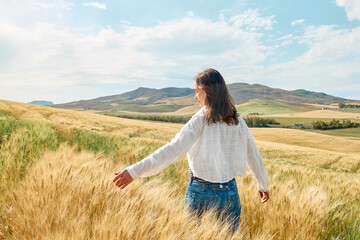 Rear view woman walking in golden wheat field in hot summer sun and blue sky with white clouds with mountains hill landscape in background. - Powered by Adobe