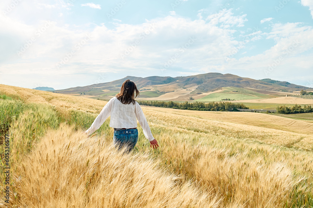 Wall mural rear view woman walking in golden wheat field in hot summer sun and blue sky with white clouds with 