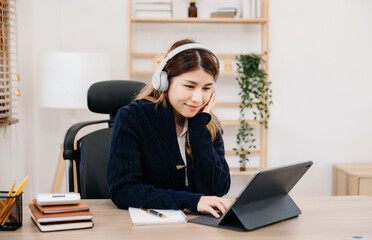 Attractive happy Asian student studying at the college library, sitting at the desk, using a laptop computer, tablet and headphones having a video chat..