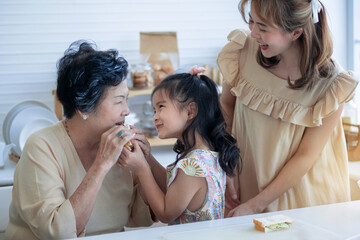 Little girl is feeding the sandwich she made for grandma, and her mother standing nearby, old woman smiled happily with her granddaughter, happy family concept