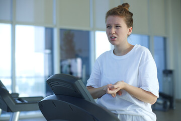 Tired exhausted young woman is training workout, running on treadmill in gym holding her head with...