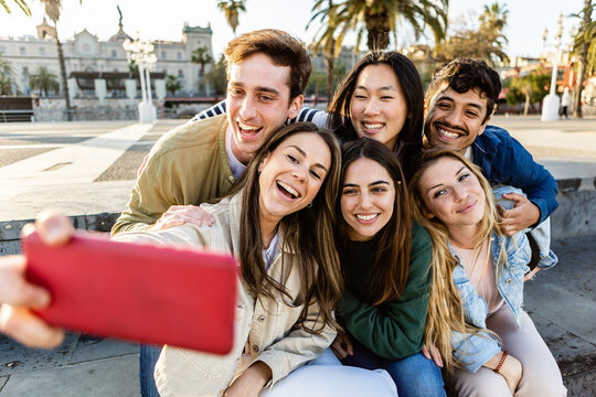 Happy young group of people taking selfies with mobile phone in city street. Teenage boys and girls having fun together enjoying day off at sunset. Friendship, youth and gen z people concept.