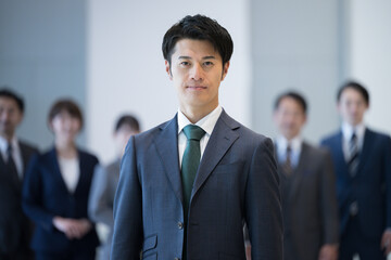 Fototapeta 若い社長や社員のイメージ　逆光の中カッコいいスーツの男性の正面のアップ　背景にビジネスマン obraz
