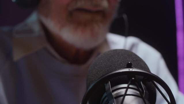 The old man creates a podcast in a professional studio. Senior male does online streaming interviews. Elderly man blogger speaking in microphone or make vlog. Recording audio content for social media