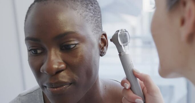 Happy diverse female patient and doctor, checking her ear with otoscope in hospital, in slow motion