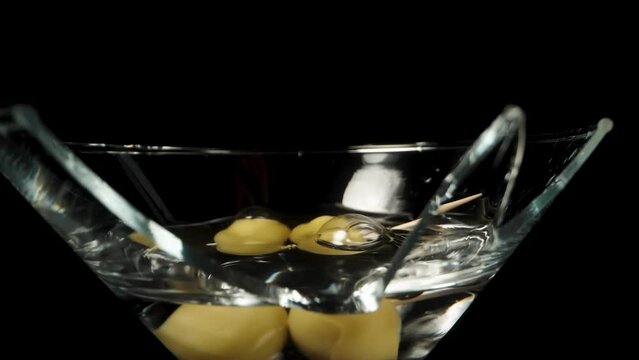 In the broken martini cocktail glass, olives on a toothpick are falling, in slow motion, dolly slider.